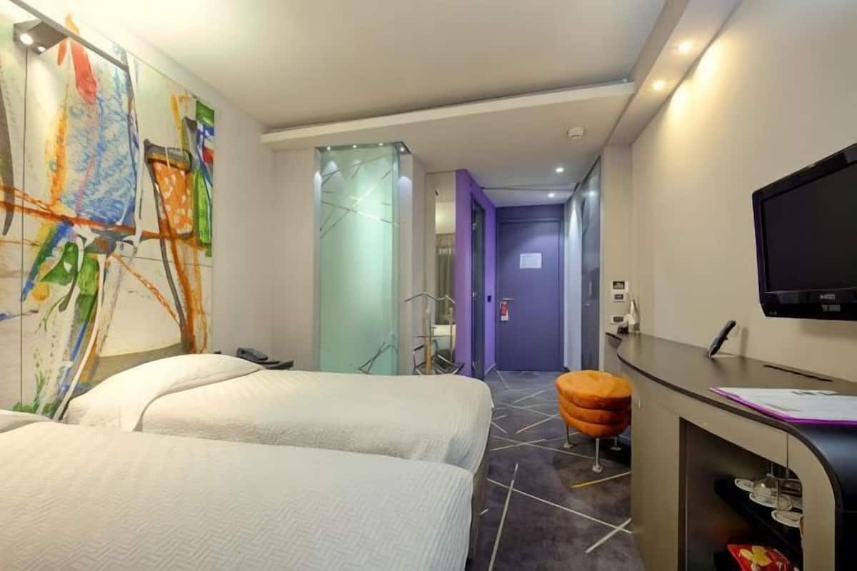 A hotel room with two beds, a desk with a computer, a TV, and a glass partition wall.