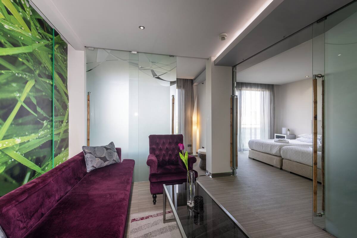 A modern hotel room with a glass partition, a purple armchair, a small table with a vase, and a bed with a white comforter.