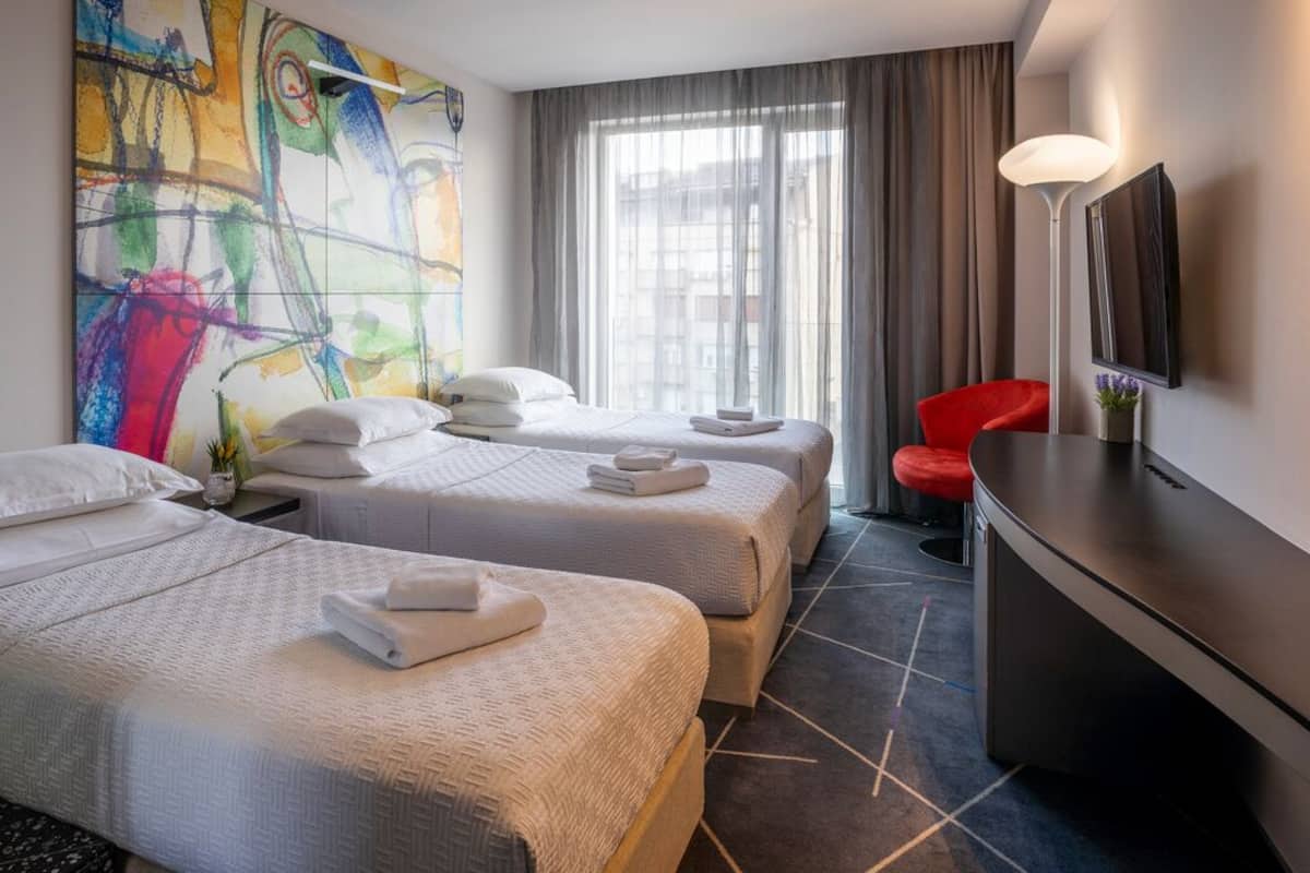 A hotel room with two beds, a desk, a red chair, a large abstract painting, and a TV on the wall.