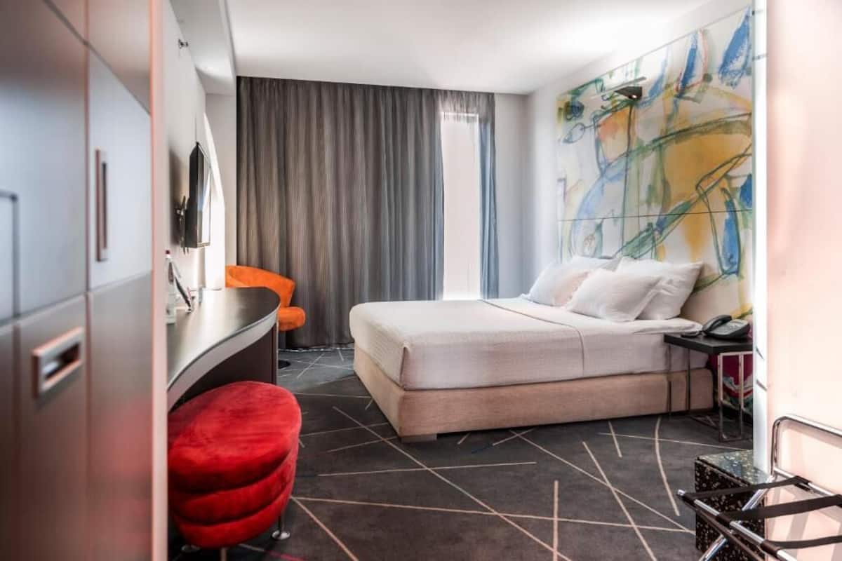 A hotel room with a bed, a desk, a red chair, a TV, and a large abstract painting on the wall.