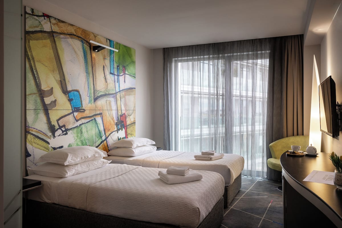 A hotel room with two beds, a large abstract painting on the wall, a desk with a lamp, and a view of the city through sheer curtains.