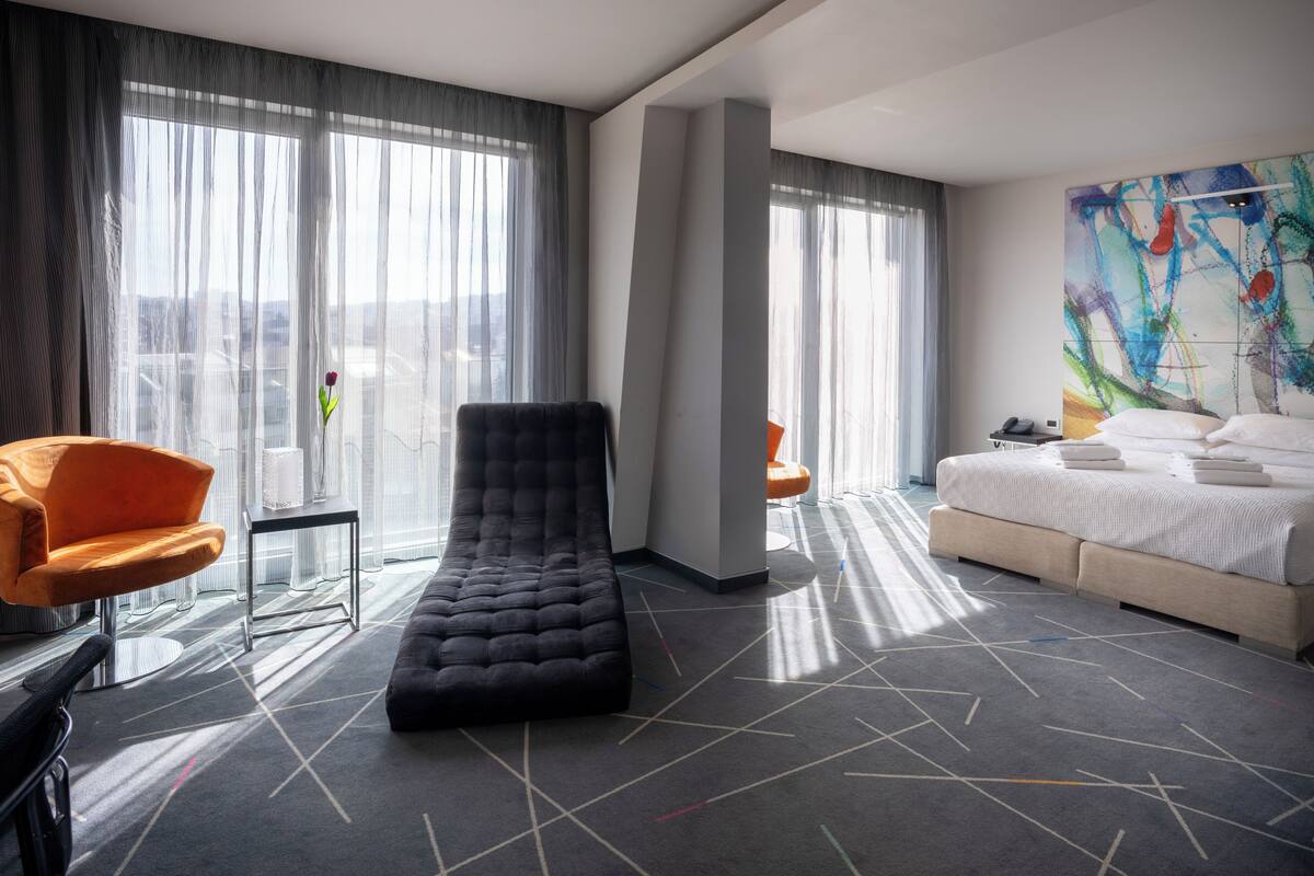 A modern hotel room with a large window, a bed, a lounge chair, and a colorful abstract painting.