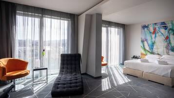 Una habitación de hotel moderna con un ventanal grande, una cama, un sillón de descanso y un cuadro abstracto de colores.