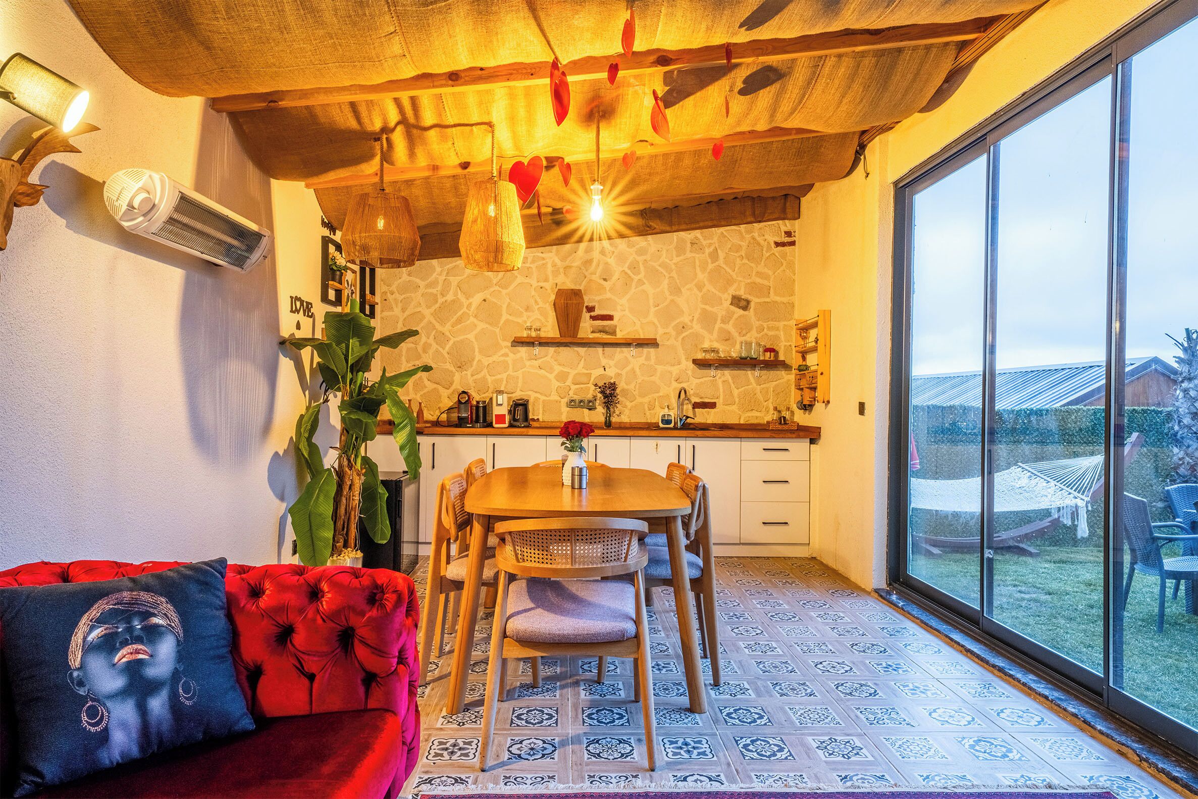A cozy living area with a red sofa, a dining table with chairs, a large window with a view of the outdoors, and a stone wall.