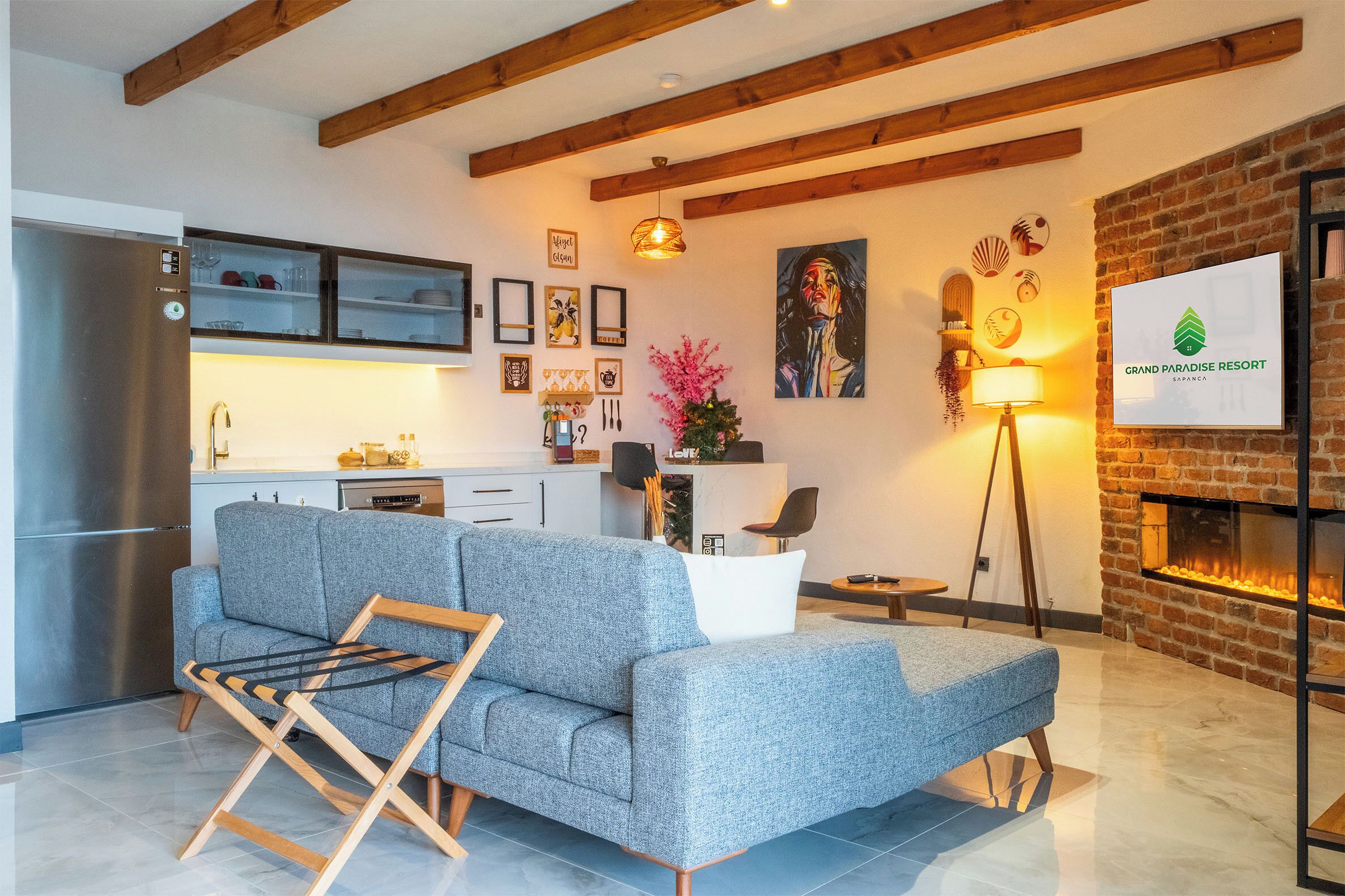 Ein modernes Wohnzimmer mit grauem Sofa, Holzbalken, Kamin und Küchenzeile mit Kühlschrank und Schränken.