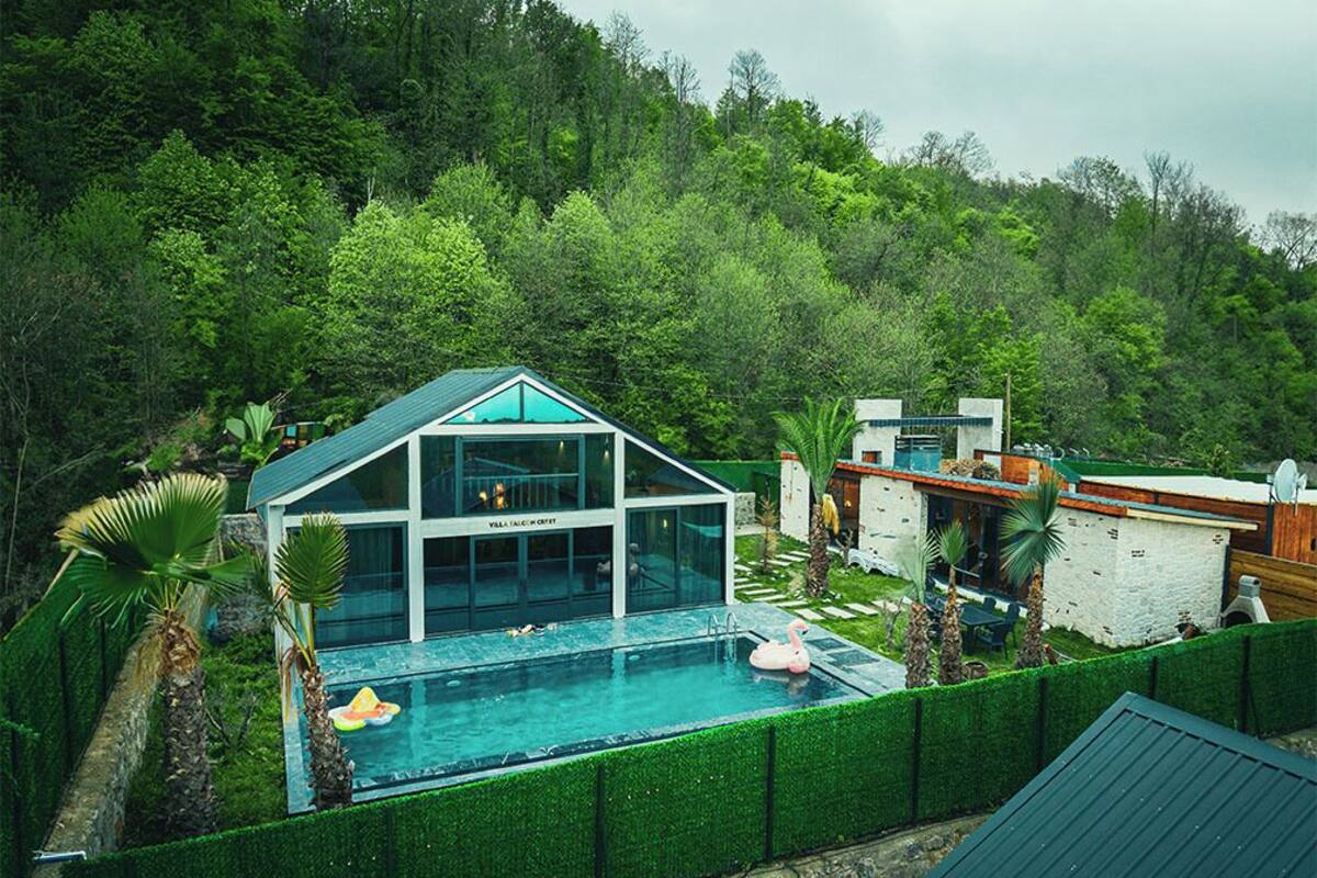 Ein modernes Haus mit einem von Glas umgebenen Pool, umgeben von üppiger Vegetation und einer hügeligen Landschaft.