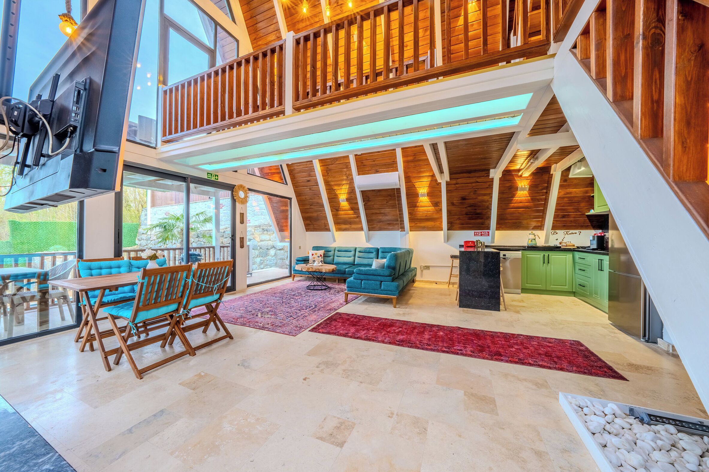 A spacious living area with a wooden ceiling, a balcony, a sofa, a dining table, and a kitchen area.