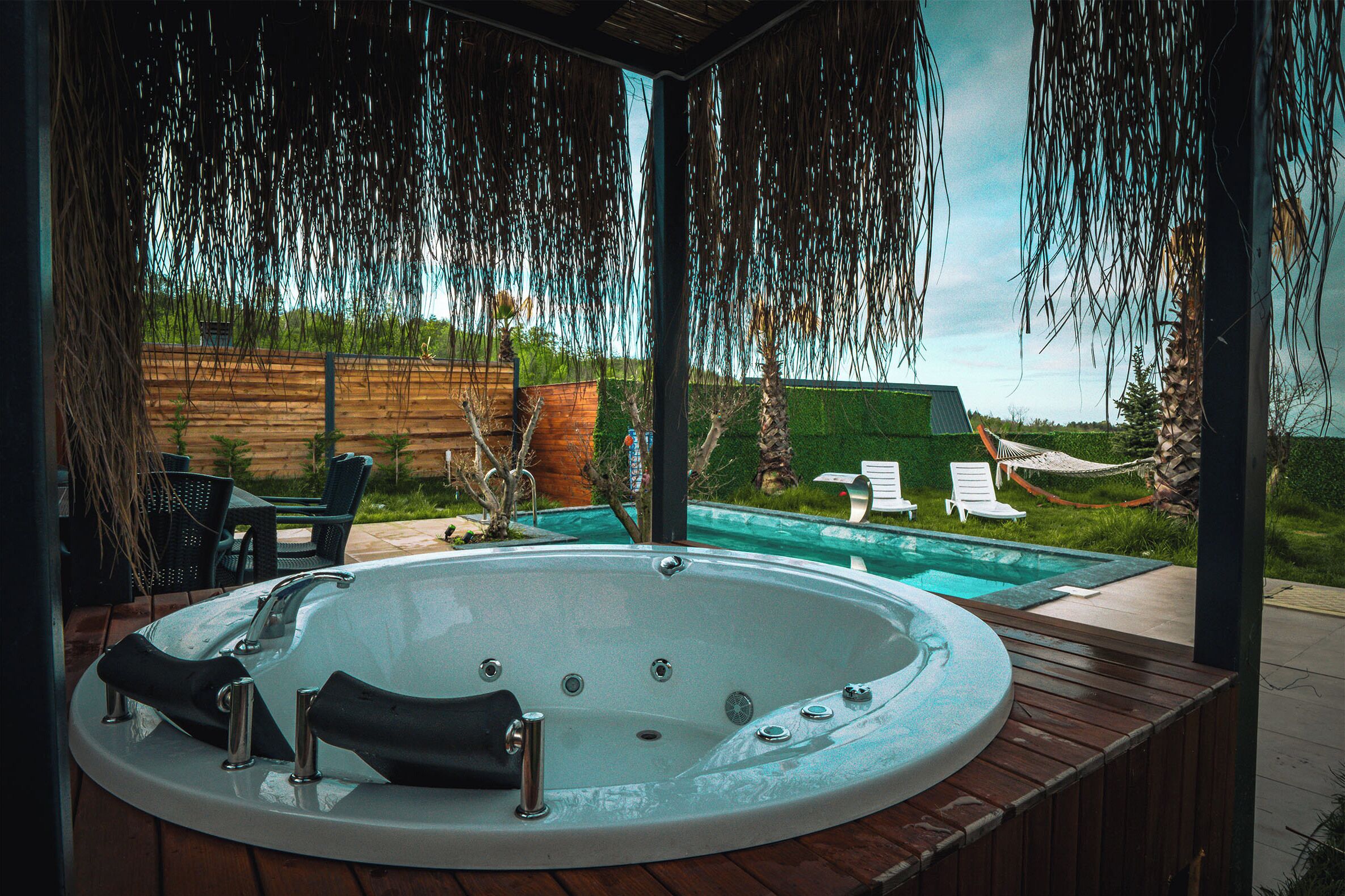 A hot tub with a wooden deck, surrounded by outdoor furniture and a view of a swimming pool.