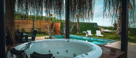 A hot tub with a wooden deck, surrounded by outdoor furniture and a view of a swimming pool.