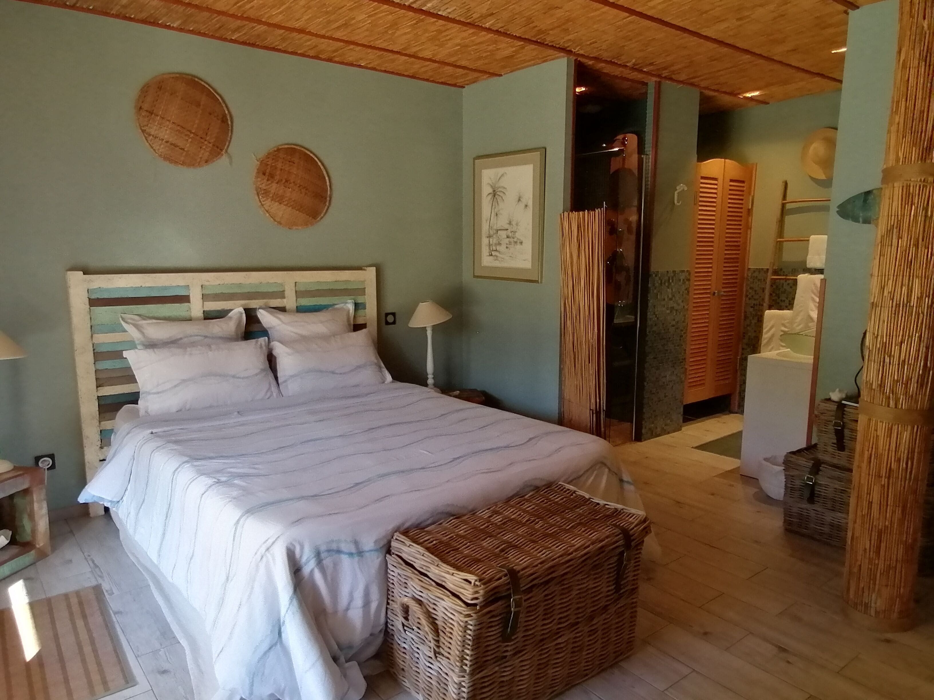 Quarto com cama, mesa de cabeceira, abajur, cesto de vime e porta que dá para outro cômodo.