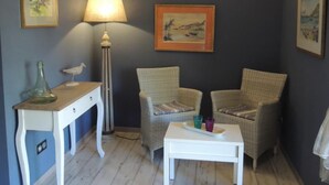 Ein Zimmer mit zwei Korbstühlen, einem kleinen weißen Tisch, einer Lampe und gerahmten Bildern an der Wand.
