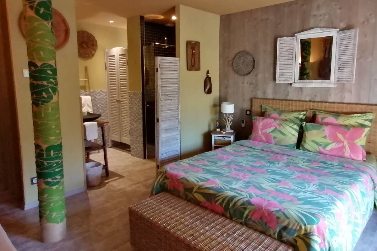 Una camera da letto con un letto, un comodino, una panca in vimini, uno specchio e una porta che conduce al bagno.