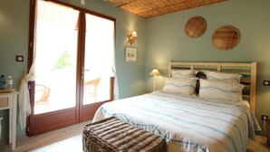 Ein Schlafzimmer mit einem Bett, einem Korbsitzhocker, einem Holz-Kopfteil, einem Fenster mit Jalousien und einer Wandlampe.