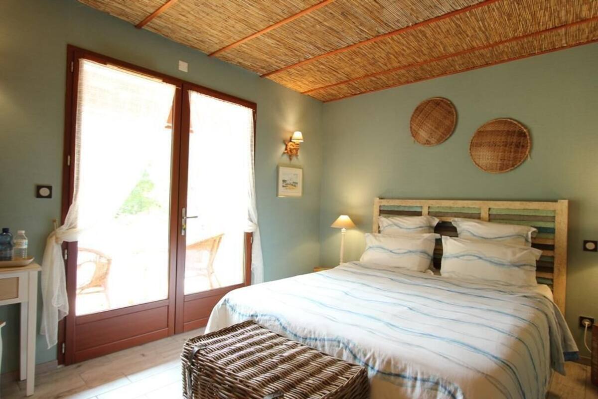 Una camera da letto con un letto, un poggiapiedi in vimini, una testiera in legno, una finestra con persiane e una lampada a parete.