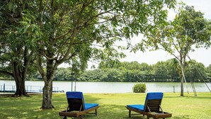 Two lounge chairs with blue cushions on a grassy area near a lake, surrounded by trees. - Waters Edge Hotel (Battaramulla)