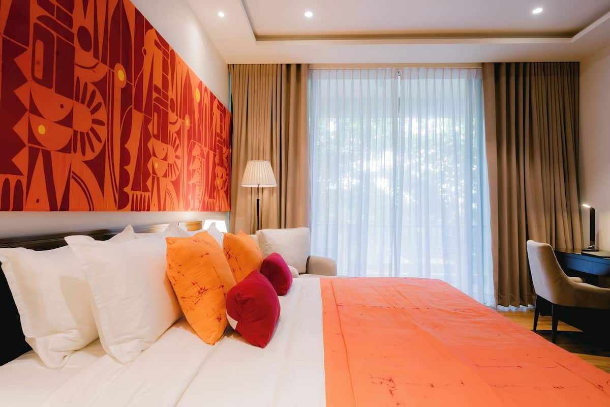 A modern hotel room with a large bed, a desk, and a view of the outdoors through sheer curtains.