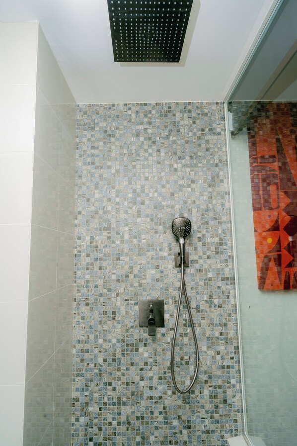 Een douchegedeelte met een betegelde muur van mozaïek, een handdouche en een in het plafond gemonteerde regendouche.