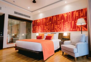A modern hotel room with a large bed, a nightstand with a lamp, a seating area, and a large abstract wall art piece. - Waters Edge Hotel (Battaramulla)