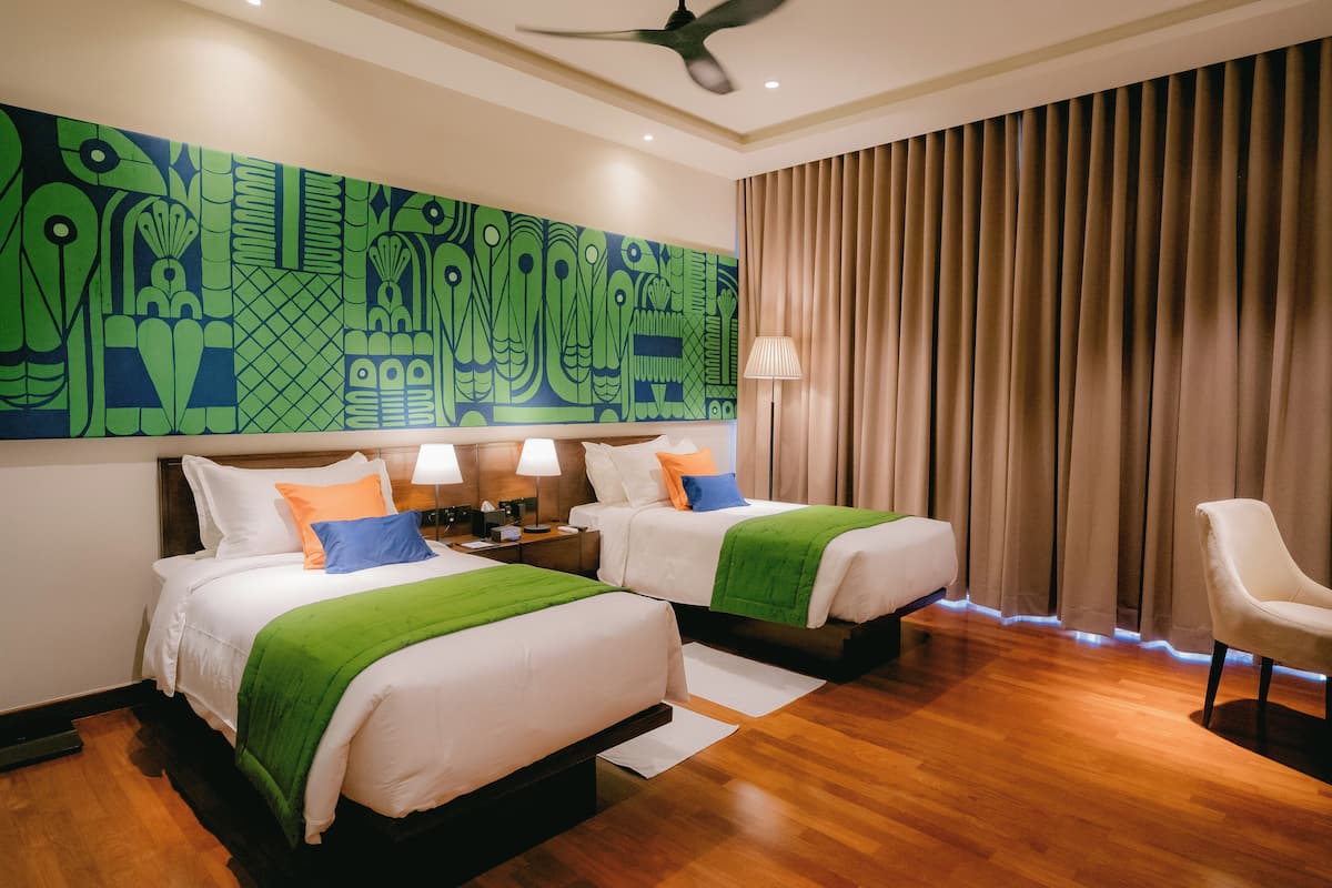 A hotel room with two beds, a ceiling fan, a large abstract painting on the wall, a lamp, and a chair.