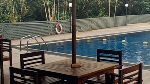 A poolside area with a table and chairs under a large umbrella, a pool with floating barriers, and a building in the background. - Waters Edge Hotel (Battaramulla)