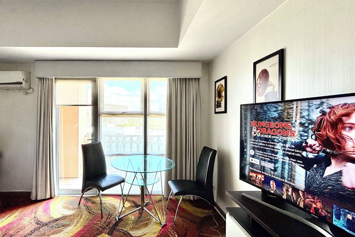 Ett modernt hotellrum med en stor TV som visar en filmplakat, ett glasbord med stolar, en balkong med gardiner och en dekorativ väggdekoration.