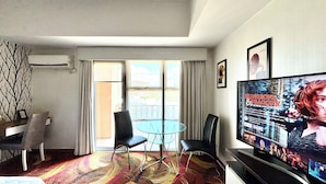 Une chambre d’hôtel moderne dotée d’un grand téléviseur affichant une affiche de film, d’une table en verre avec des chaises, d’un balcon avec des rideaux et d’un élément décoratif mural.