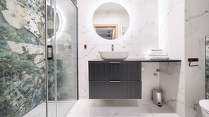 Une salle de bain moderne avec une douche à parois de verre, un miroir rond, un lavabo sur colonne et des murs en marbre.