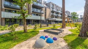 Un immeuble d’appartements moderne doté de balcons, d’un terrain de jeux avec du sable et des ballons, et d’une pelouse bien entretenue.