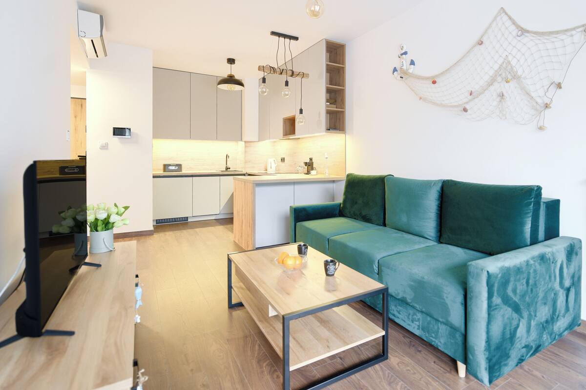 A modern living room with a teal sofa, a wooden coffee table, and a kitchen area in the background.