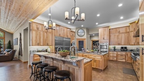 A spacious kitchen with wooden cabinets, a large island with stools, stainless steel appliances, and a chandelier.