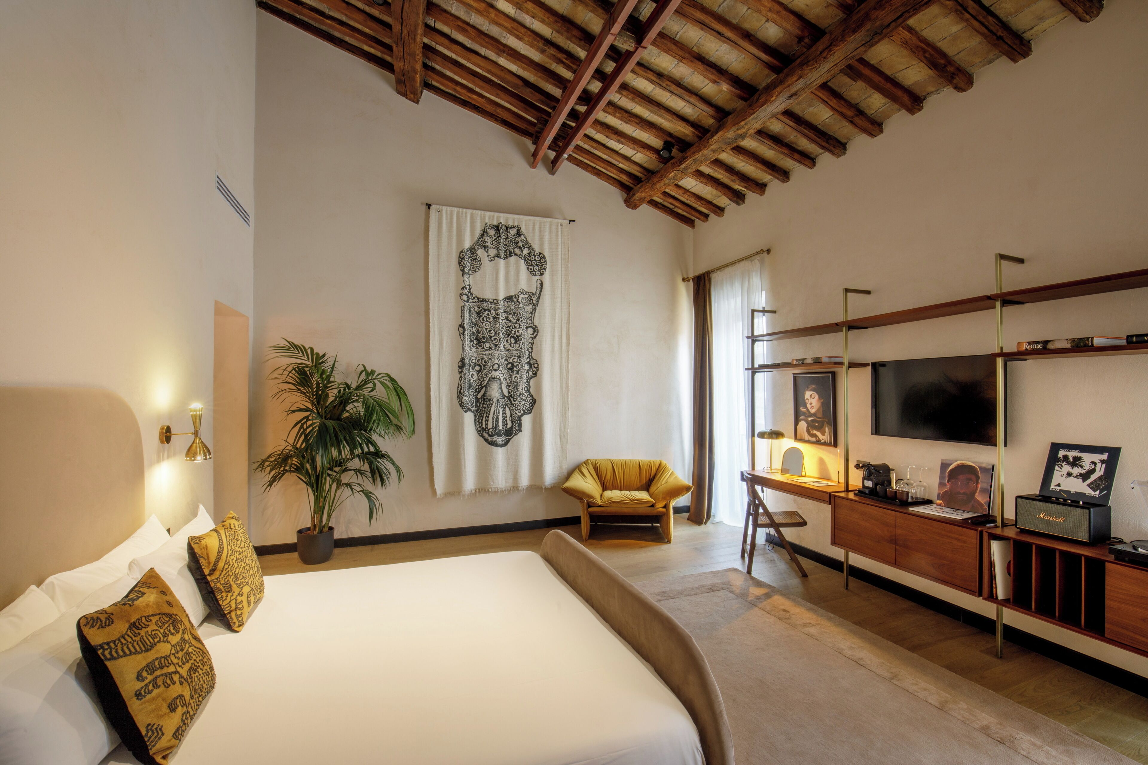 A bedroom with a large bed, a desk with a computer, a chair, a television, and a large tapestry on the wall.
