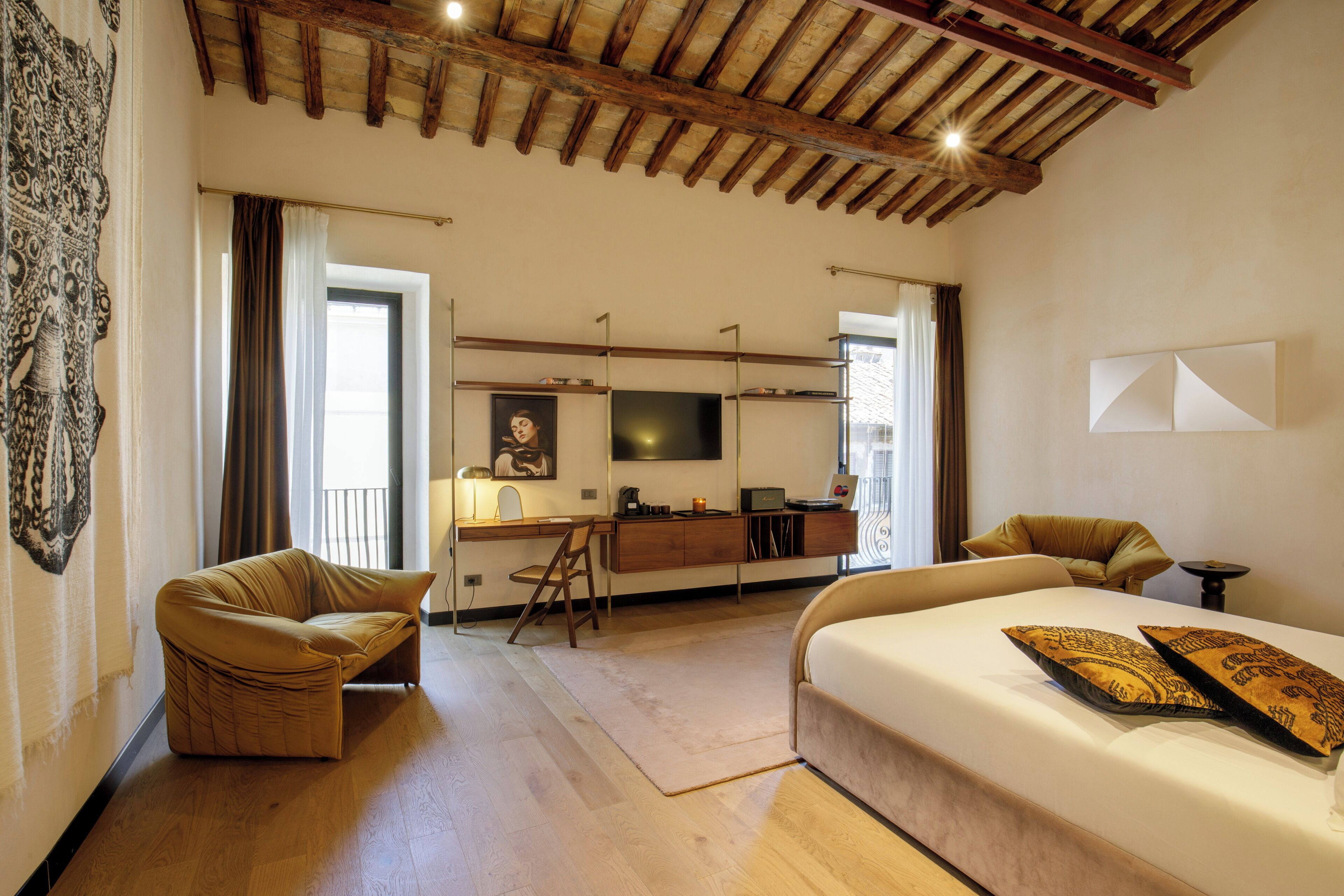 A hotel room with a bed, two armchairs, a desk with a computer, a television, and a large window with curtains.