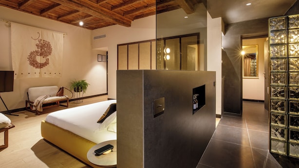 A modern hotel room with a wooden ceiling, a bed with a white mattress, a bedside table, a chair, and a large mirror. - Palazzo Serpe Suites near Colosseum (Rome)