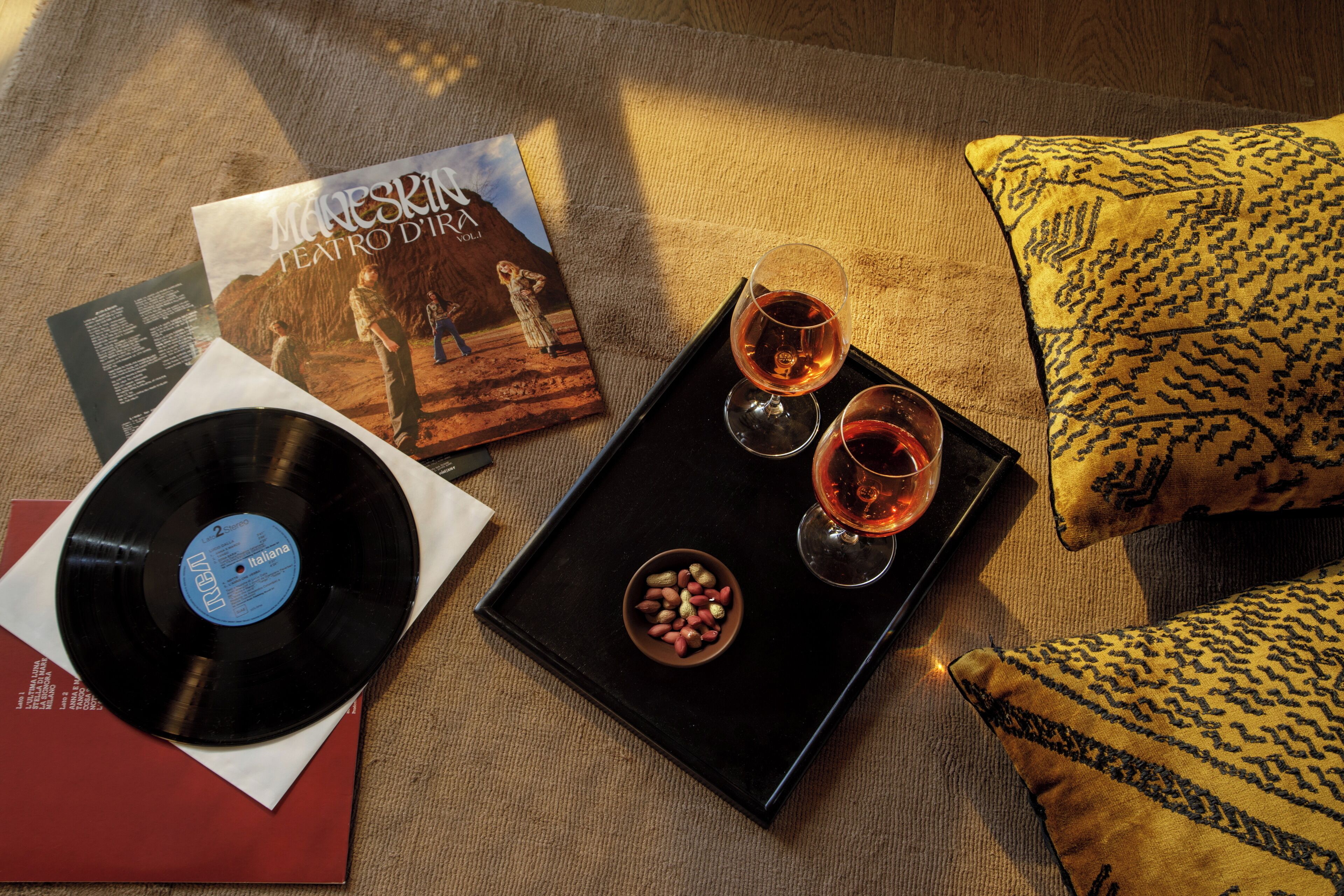 Une pièce chaleureuse avec un coussin jaune à motifs, un plateau noir sur lequel reposent deux verres de vin rouge, un disque vinyle et un magazine, le tout posé sur un tapis beige.