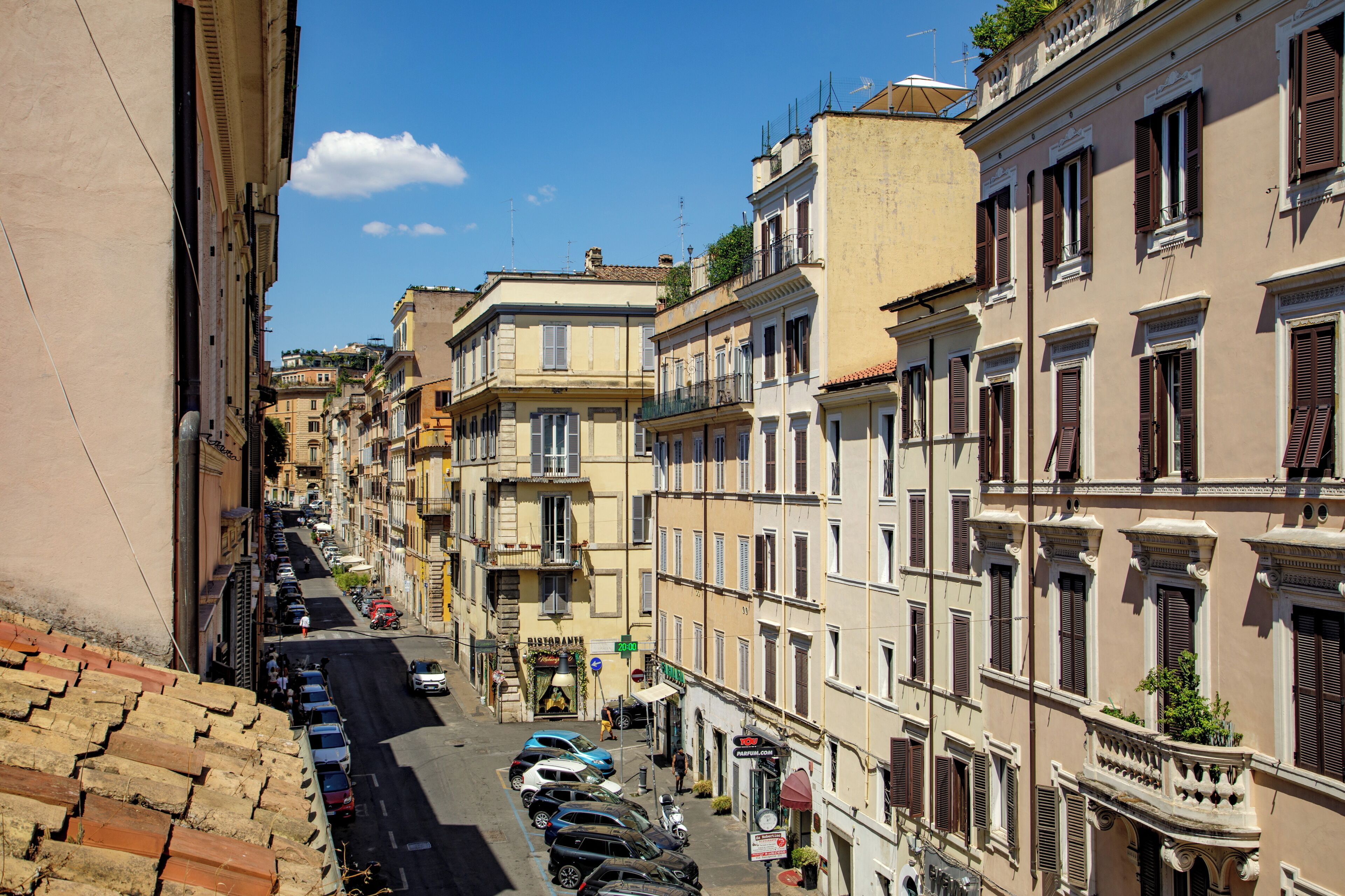 Una stretta via fiancheggiata da edifici storici, una fila di auto parcheggiate e un cielo azzurro limpido.