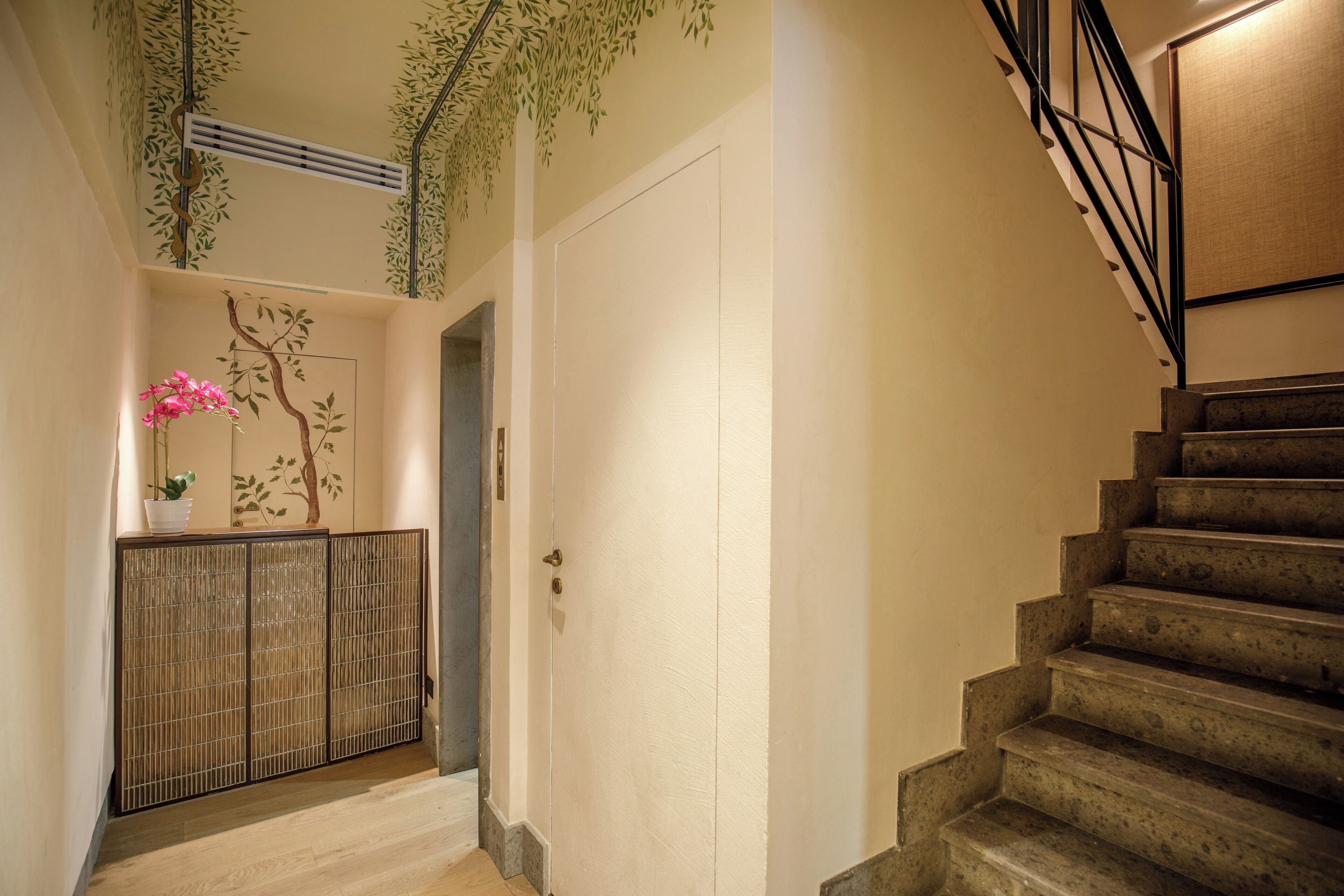 Un escalier menant à un palier où se trouvent une porte, une bouche d’aération fixée au mur et une plante décorative.