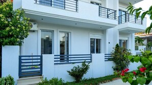 A modern two-story building with a white exterior, a balcony on the upper floor, and a small garden area with plants and flowers.