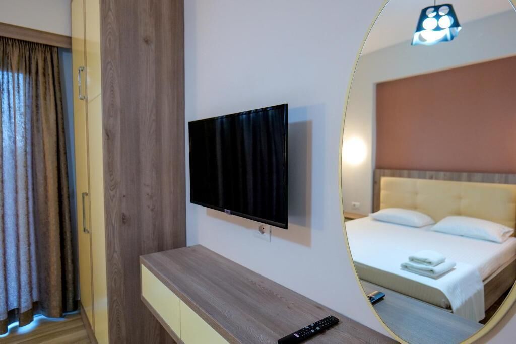 A hotel room with a wooden desk, a flat-screen TV, a bed with white linens, and a mirror reflecting the room.
