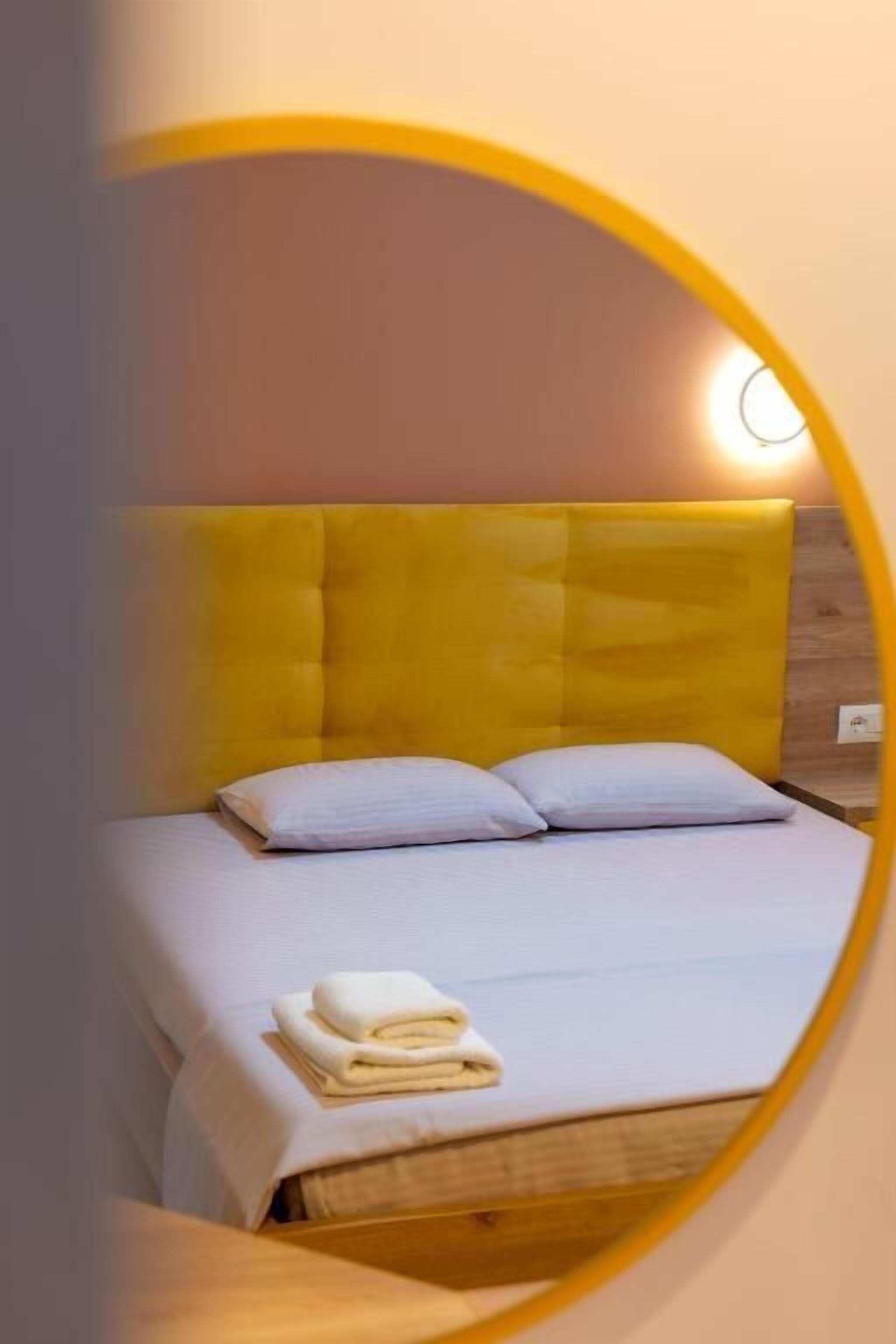 A bed with a yellow headboard and white bedding, a folded towel on the bed, and a wall-mounted light.