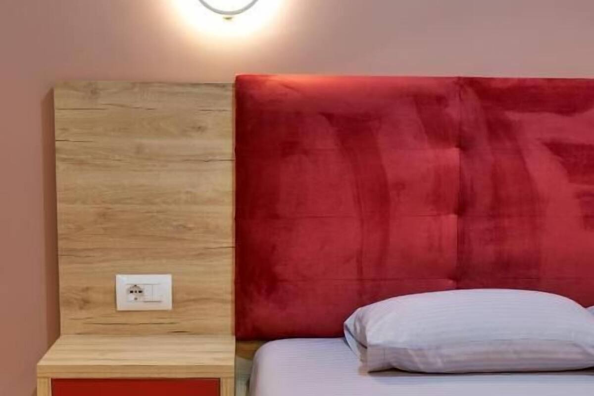 Un dormitorio con cabecera de madera, una pared de acento roja, una mesita de noche con un cajón rojo, y una cama bien hecha con almohada blanca y toallas dobladas.