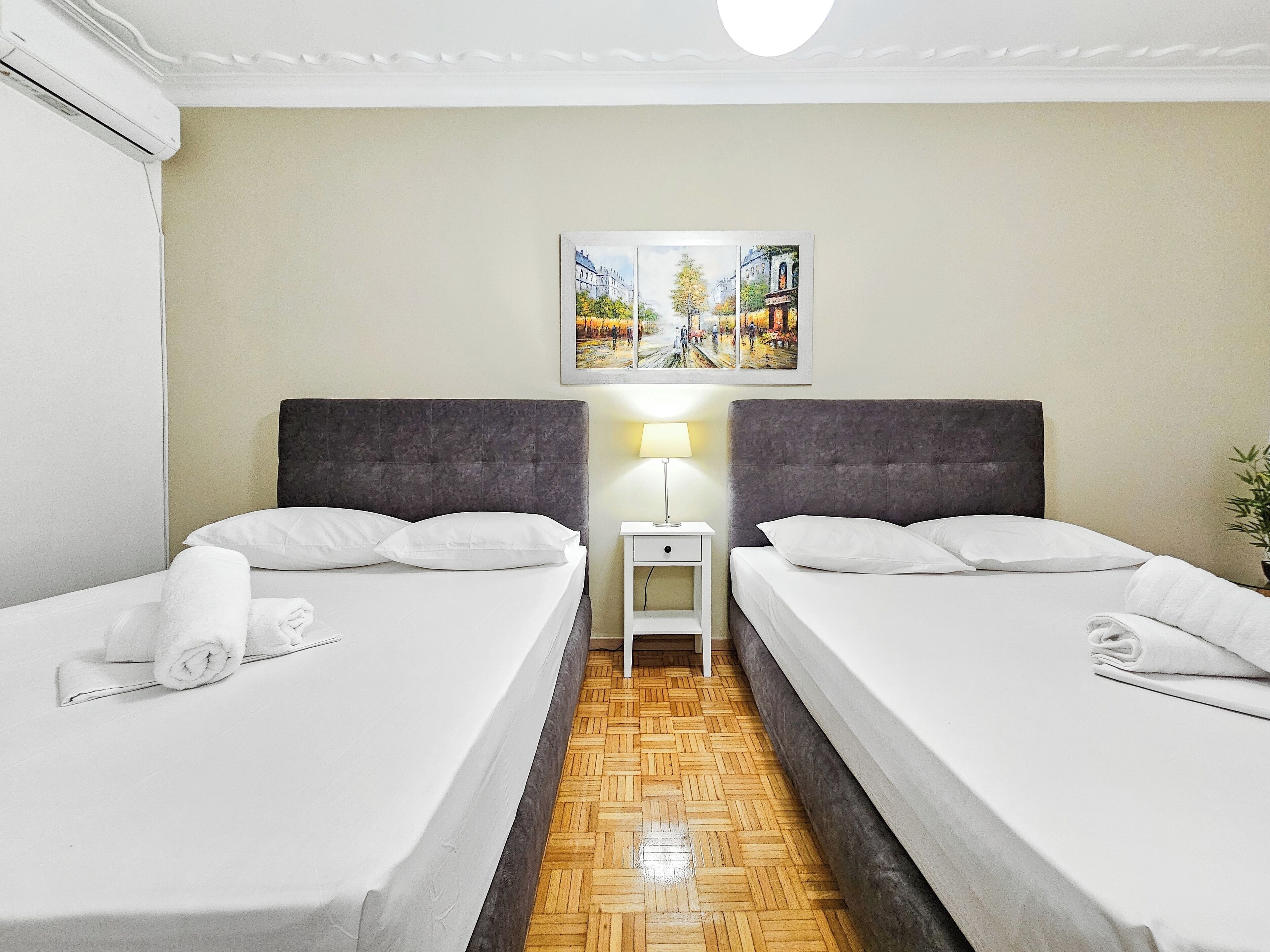 A hotel room with two beds, a nightstand with a lamp, a painting on the wall, and a wooden floor.