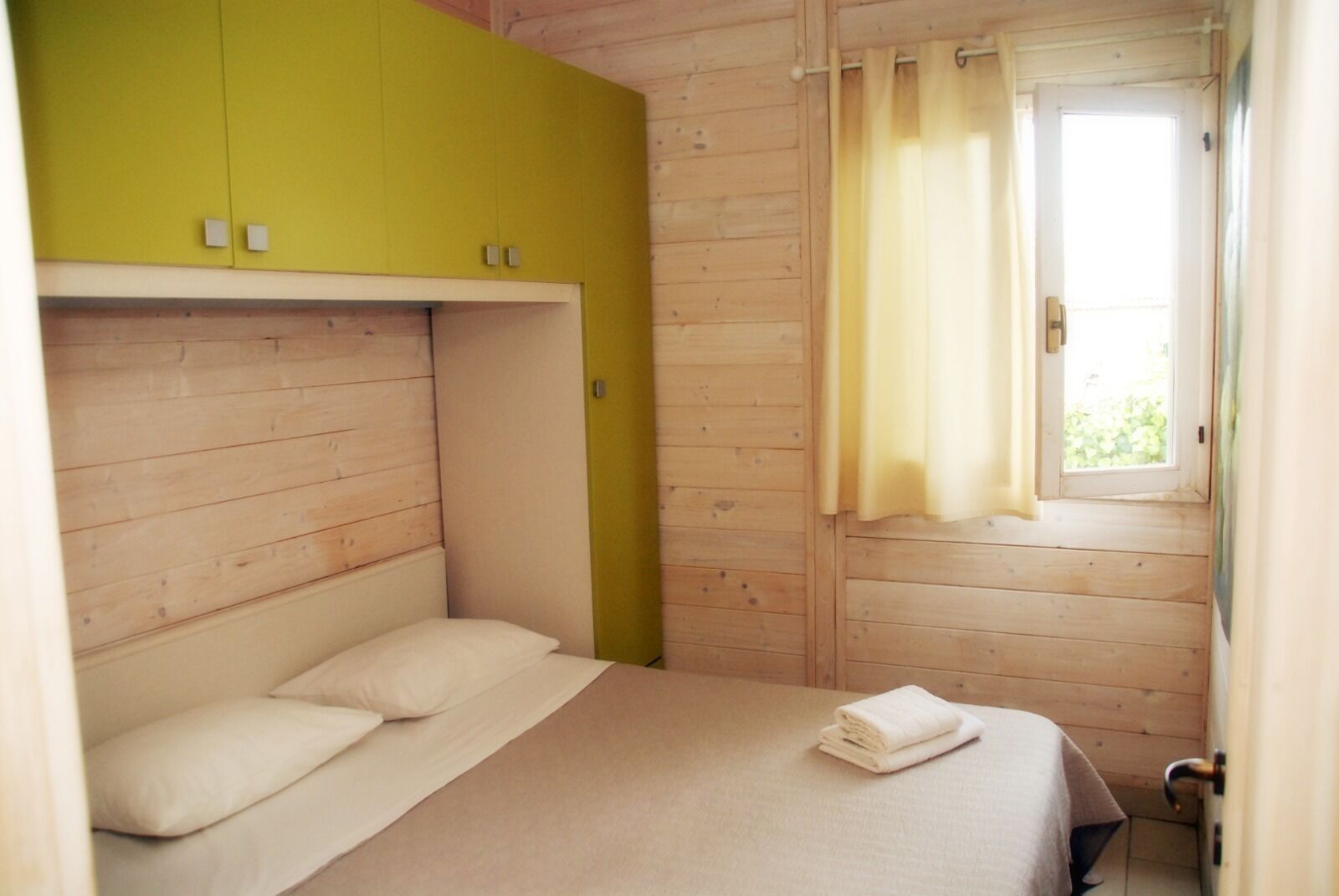 A single bed with a white mattress and pillows, a yellow wardrobe, and a window with curtains.