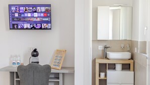 Una habitación moderna con una televisión de pantalla plana mostrando la interfaz de un servicio de streaming, un escritorio con una silla, un dispensador de agua y un menú enmarcado en la pared.
