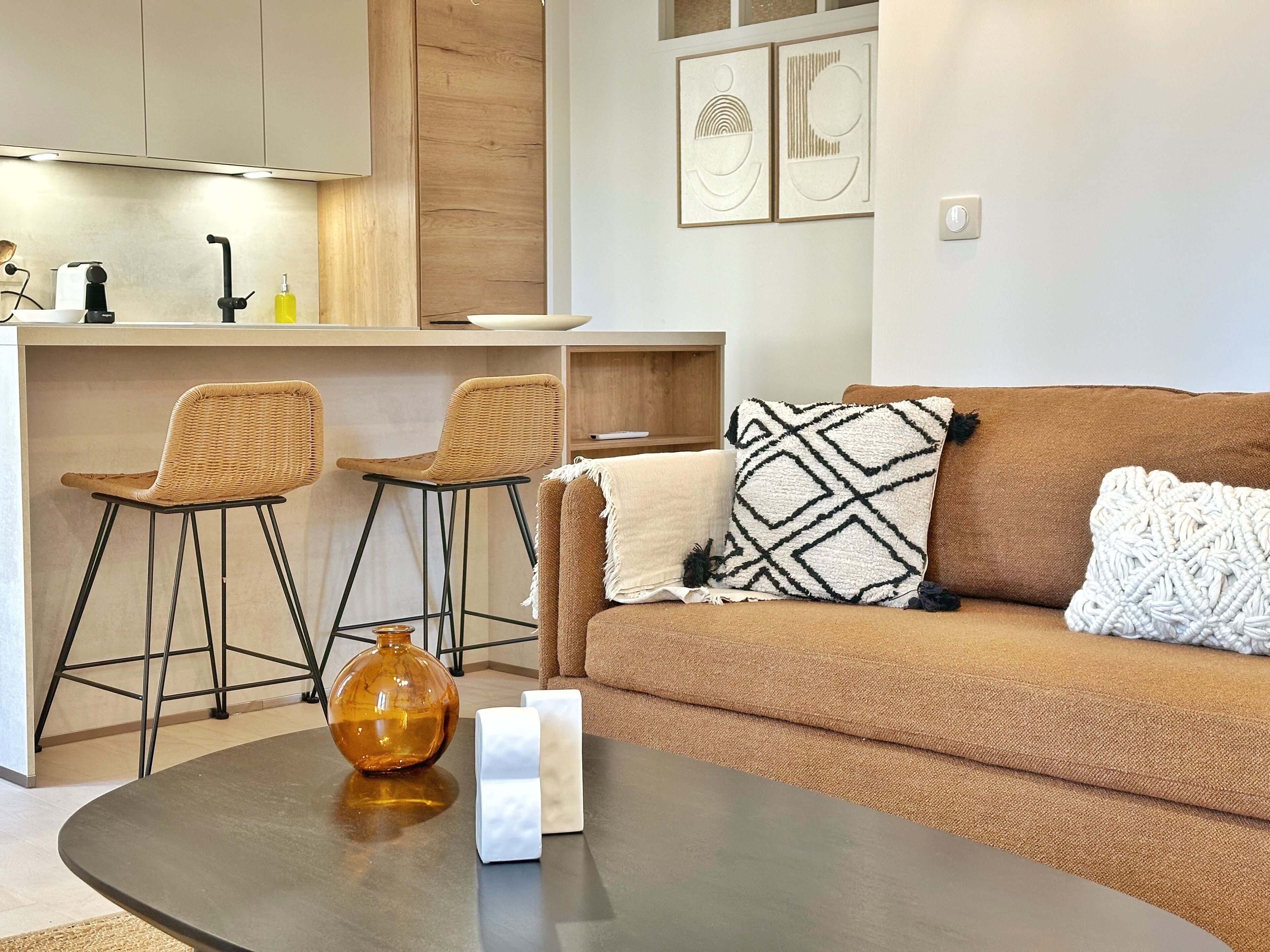 A modern living room with a brown sofa, two wicker chairs, a coffee table, and a kitchen area in the background.