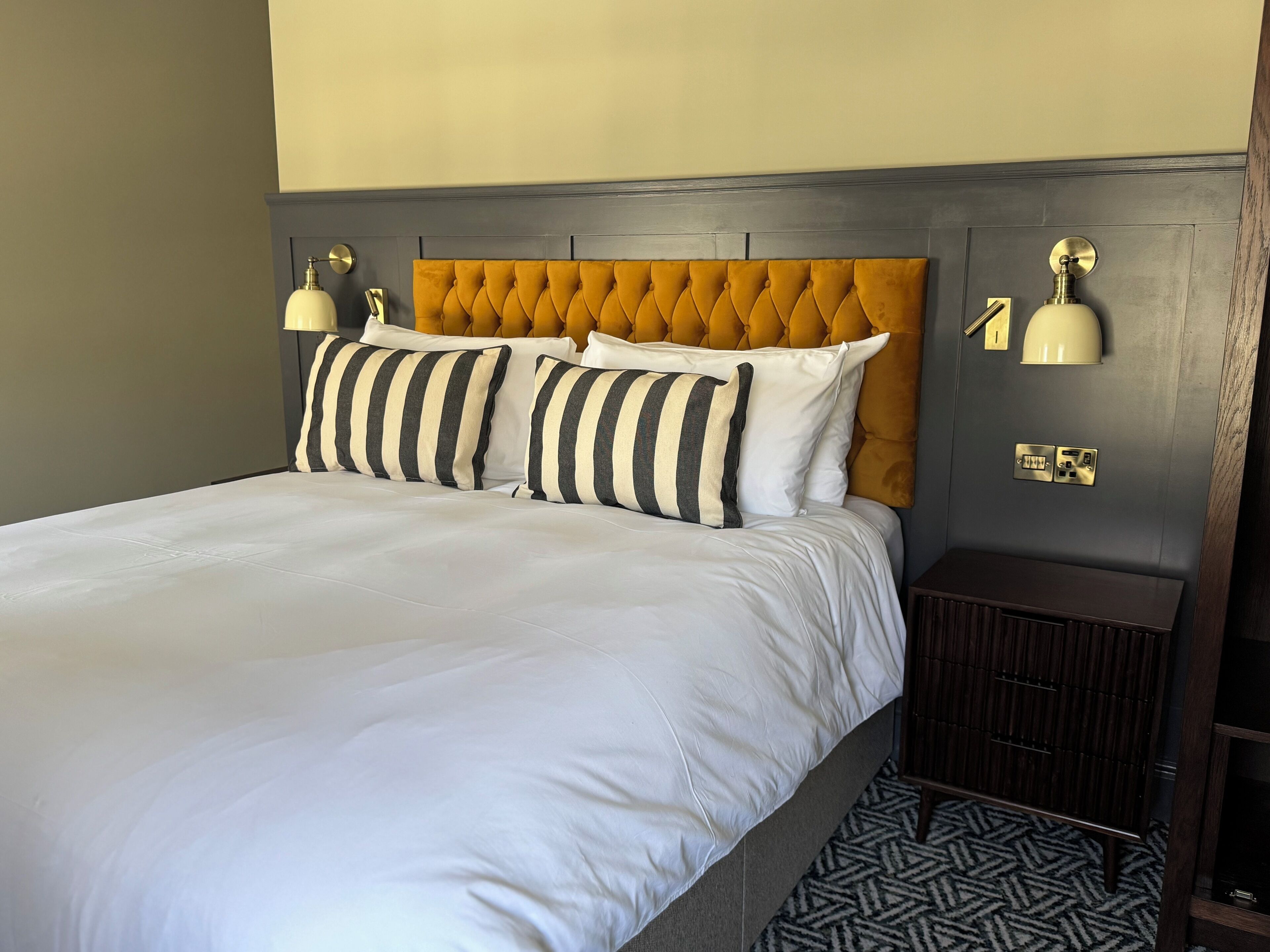 Cama com roupa de cama branca, cabeceira estofada e almofadas listradas. Há luminárias fixadas na parede e uma pequena mesa de cabeceira de madeira com um tapete estampado.