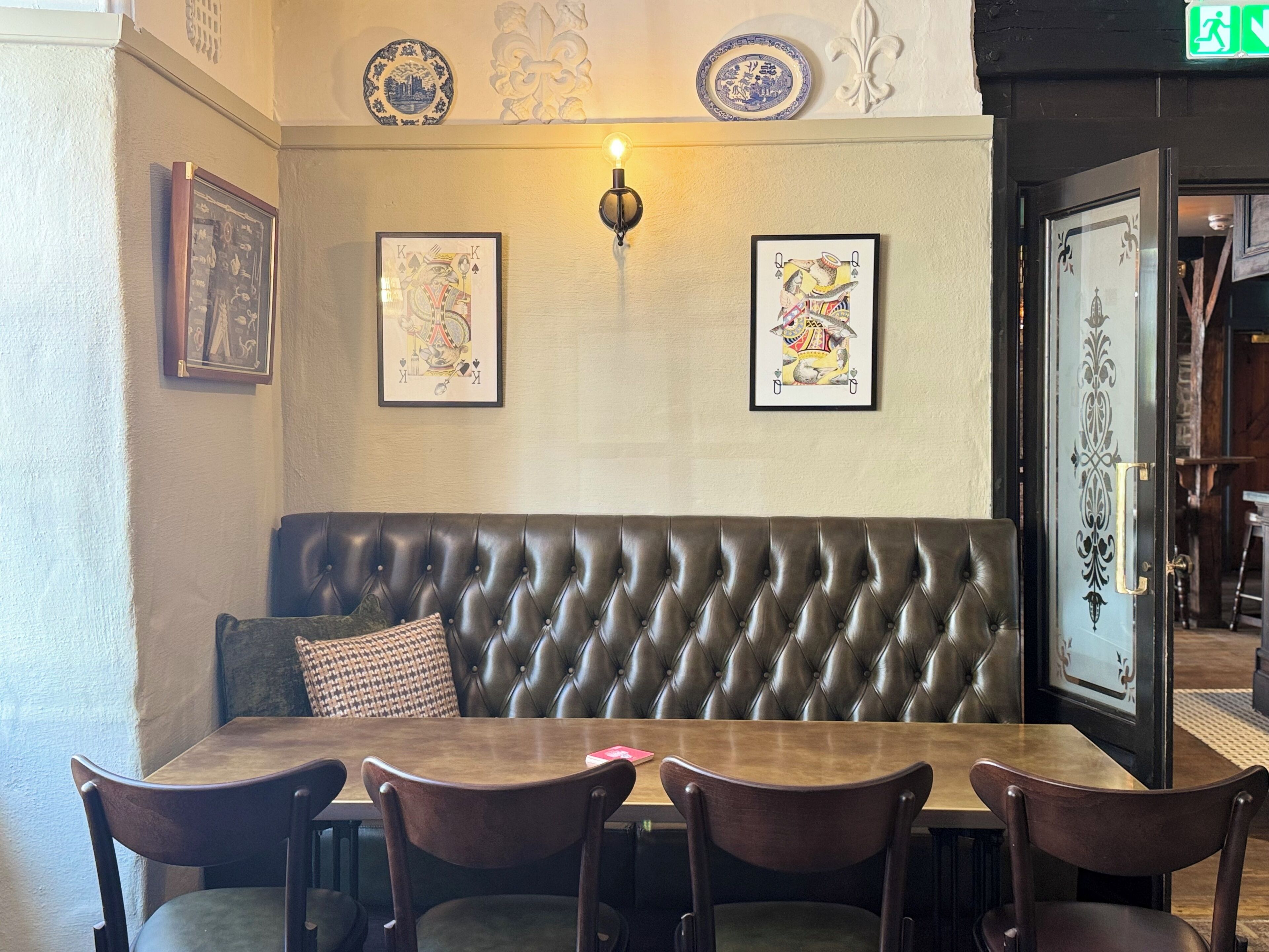 A restaurant interior with a wooden table, four chairs, a tufted leather bench, and wall-mounted artwork.