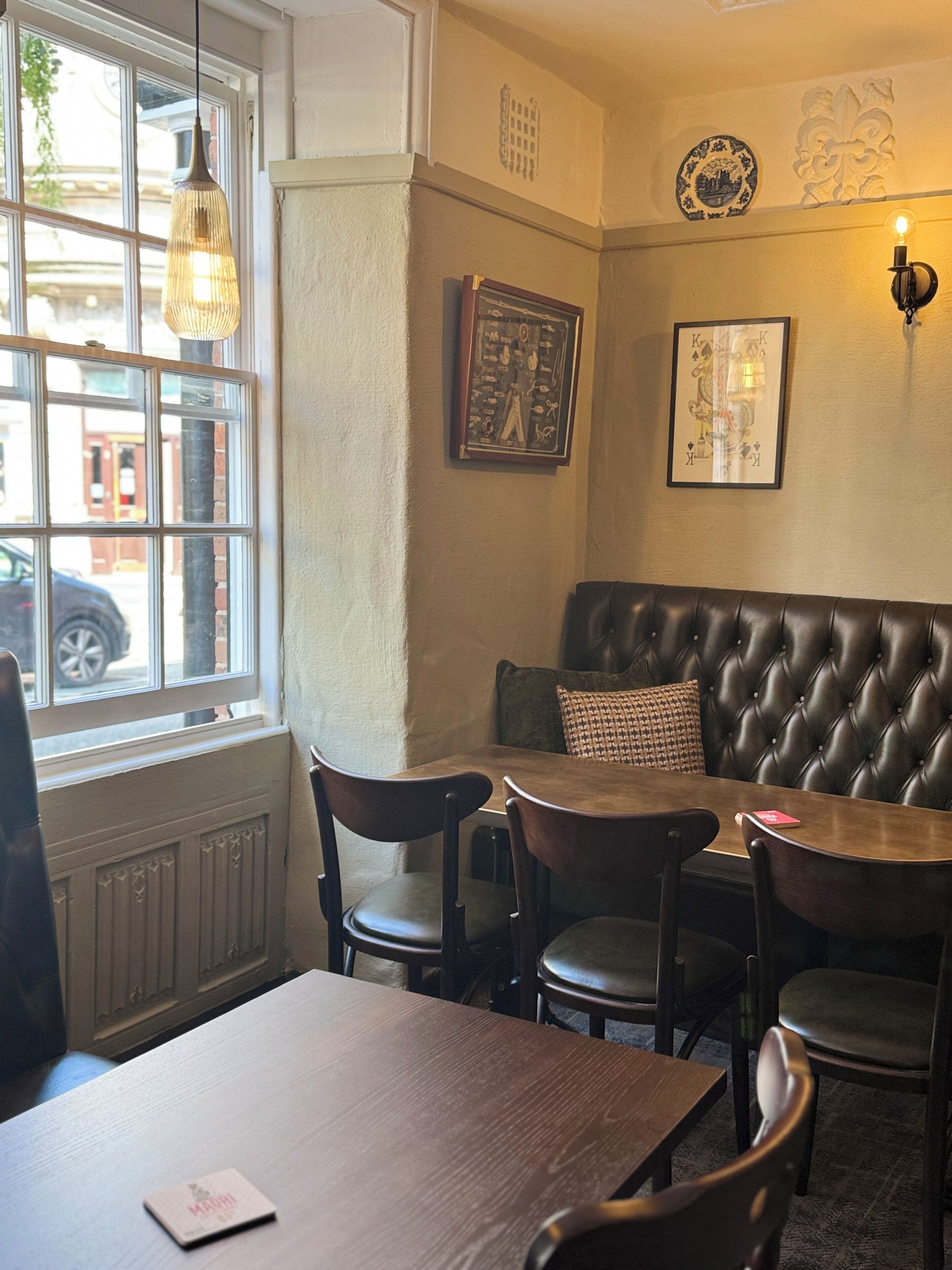 A cozy corner with a leather booth, wooden chairs, and a table. A chalkboard with writing is on the wall, and a framed playing card hangs above.