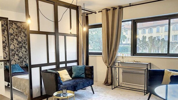 A modern living room with a sofa, a small round table, and a view of buildings through large windows. - Apartment in Biarritz (Biarritz)