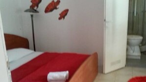 A bedroom with a bed, a red blanket, and a white pillow. There is a red rug on the floor and a white wall with a red object hanging on it. - Apartments Štrbić (Gornji Karin)