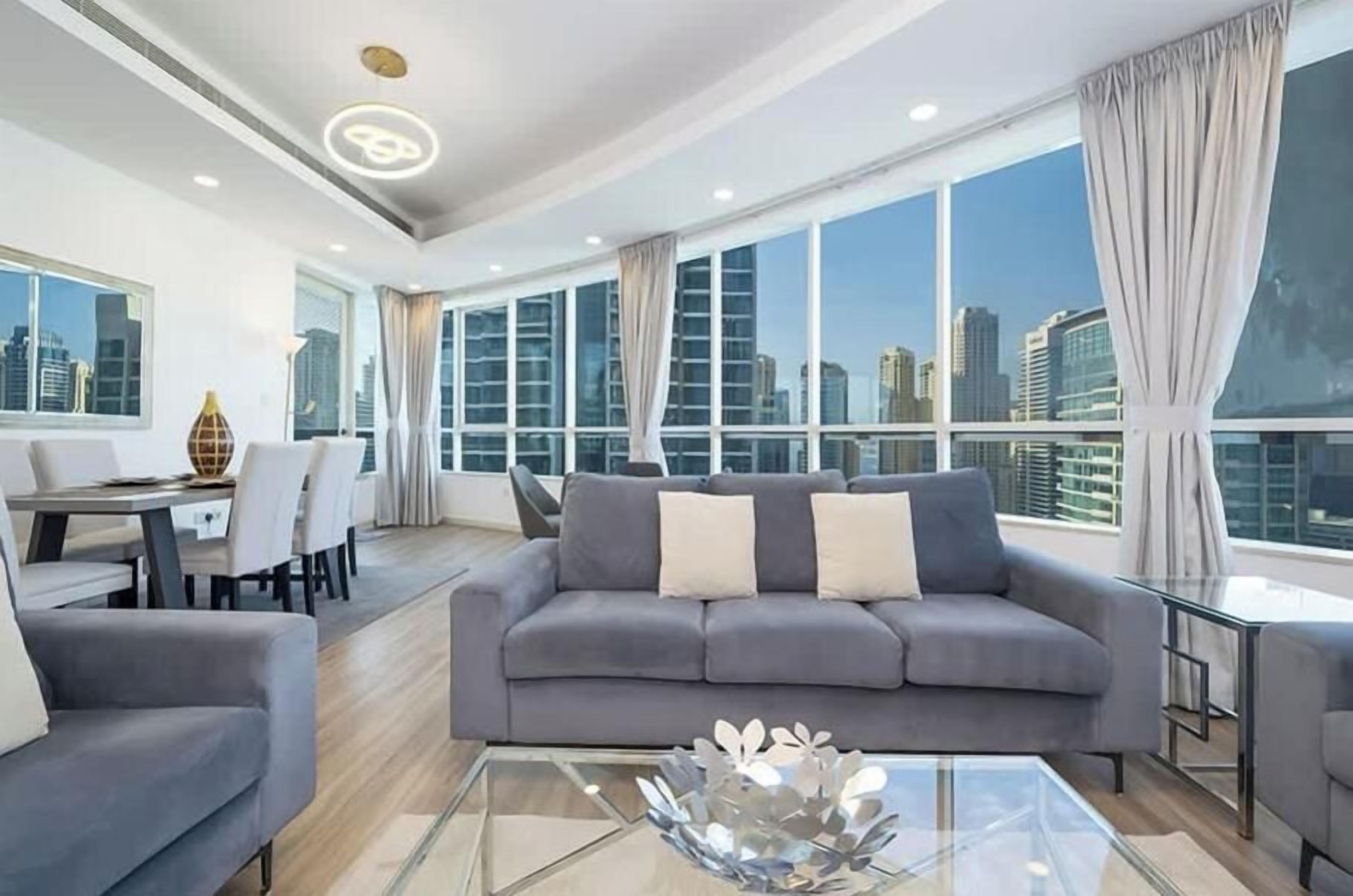 A modern living room with a gray sofa, a glass coffee table, and a city view through large windows.
